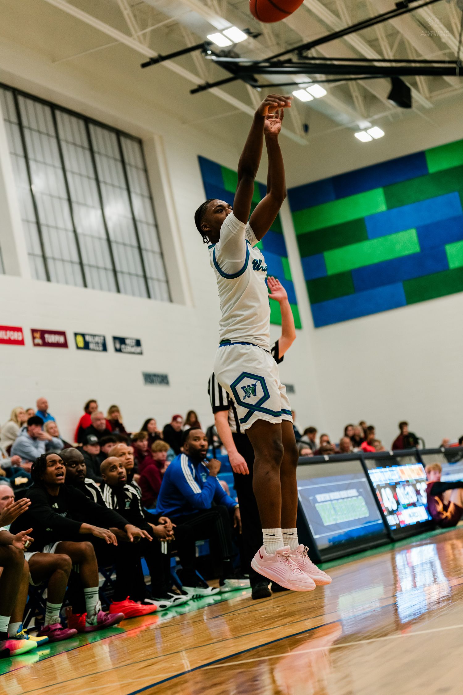 A basketball player in white uniform jumps to shoot the ball during an indoor high school basketball game.