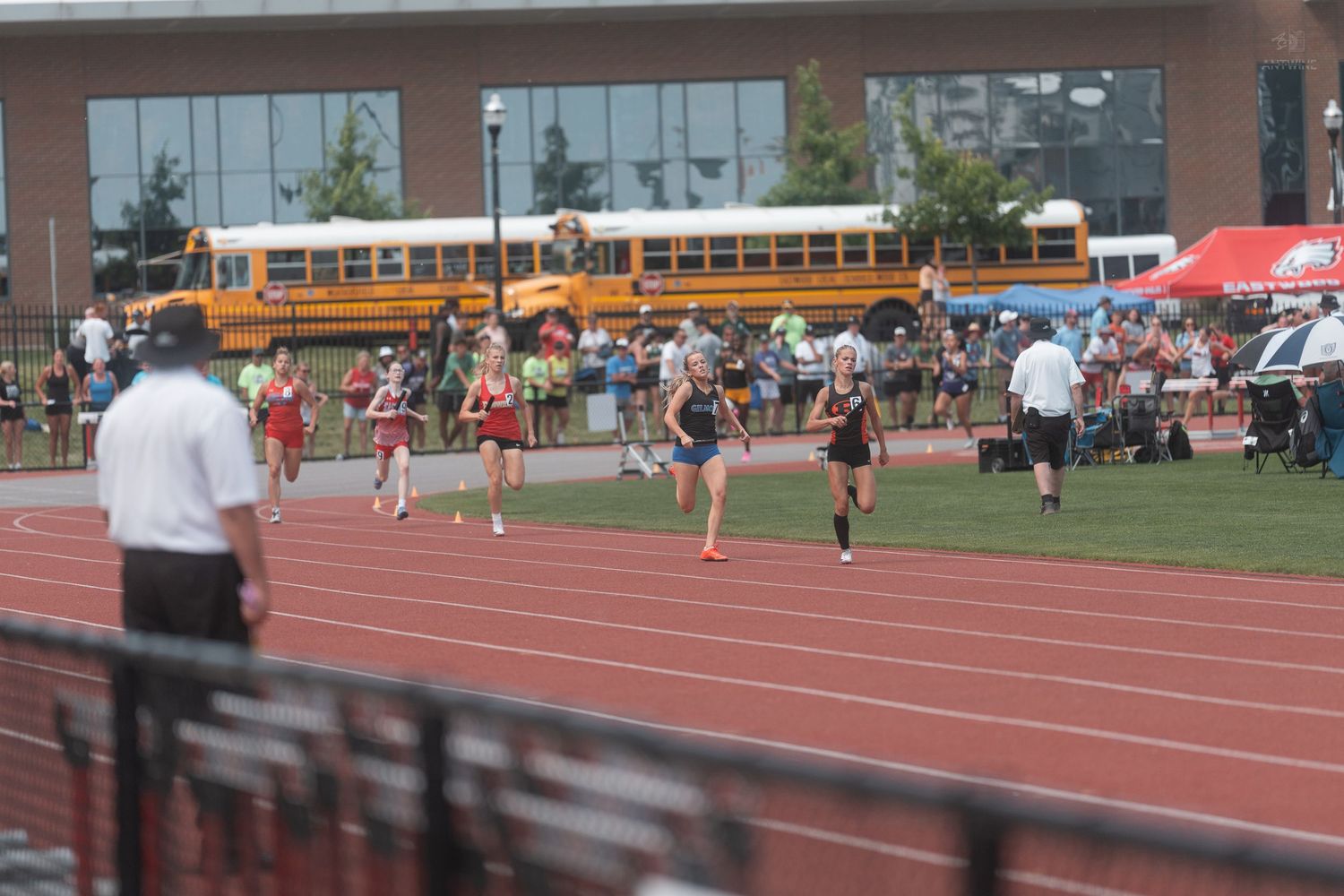 Multiple runners compete in a track race with spectators and school buses visible in the background.