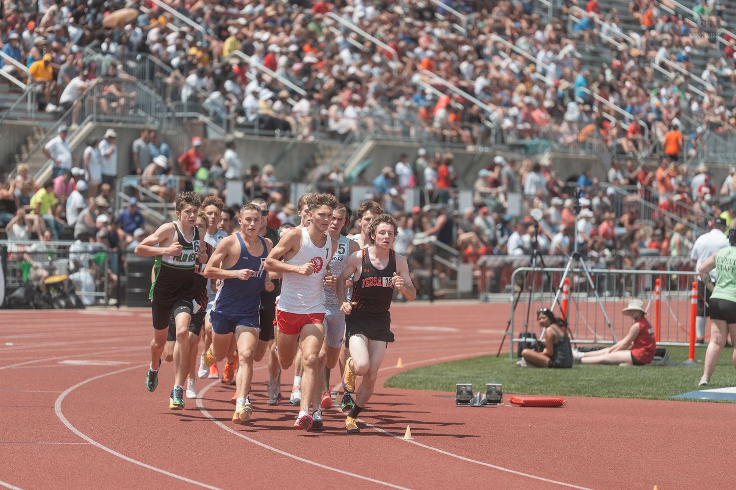 High school athletes compete in a distance running event on a red track with a large crowd in the stands.