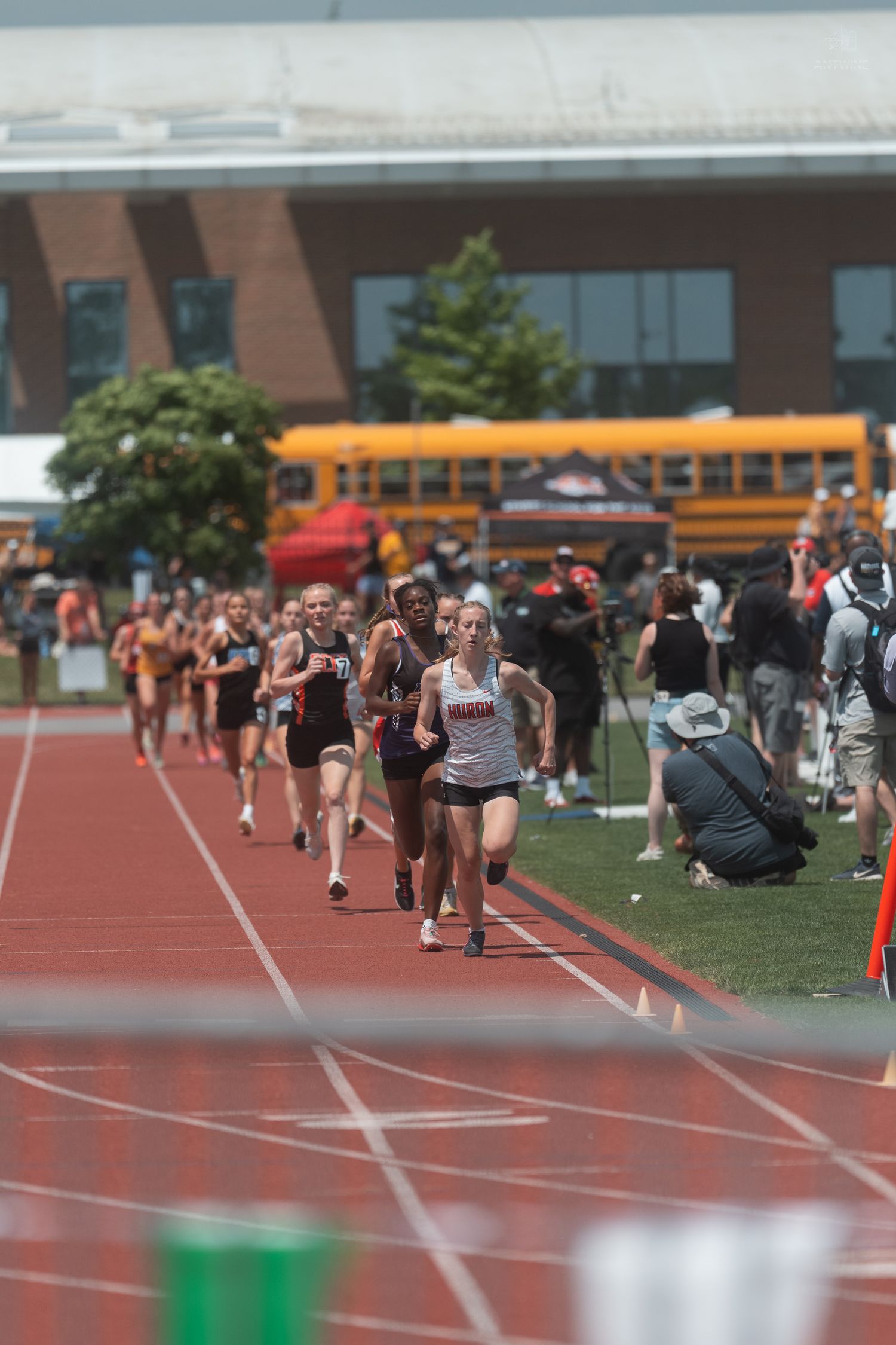 Female high school runners compete in a distance race on a red track with a school bus visible in the background.