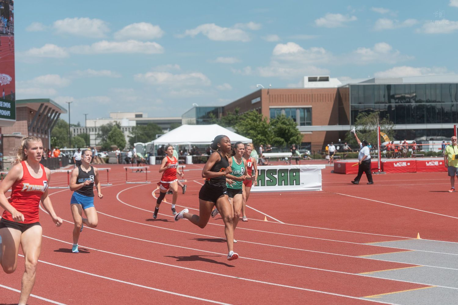 Female runners competing in an outdoor track race on a red track at what appears to be a state-level championship meet.