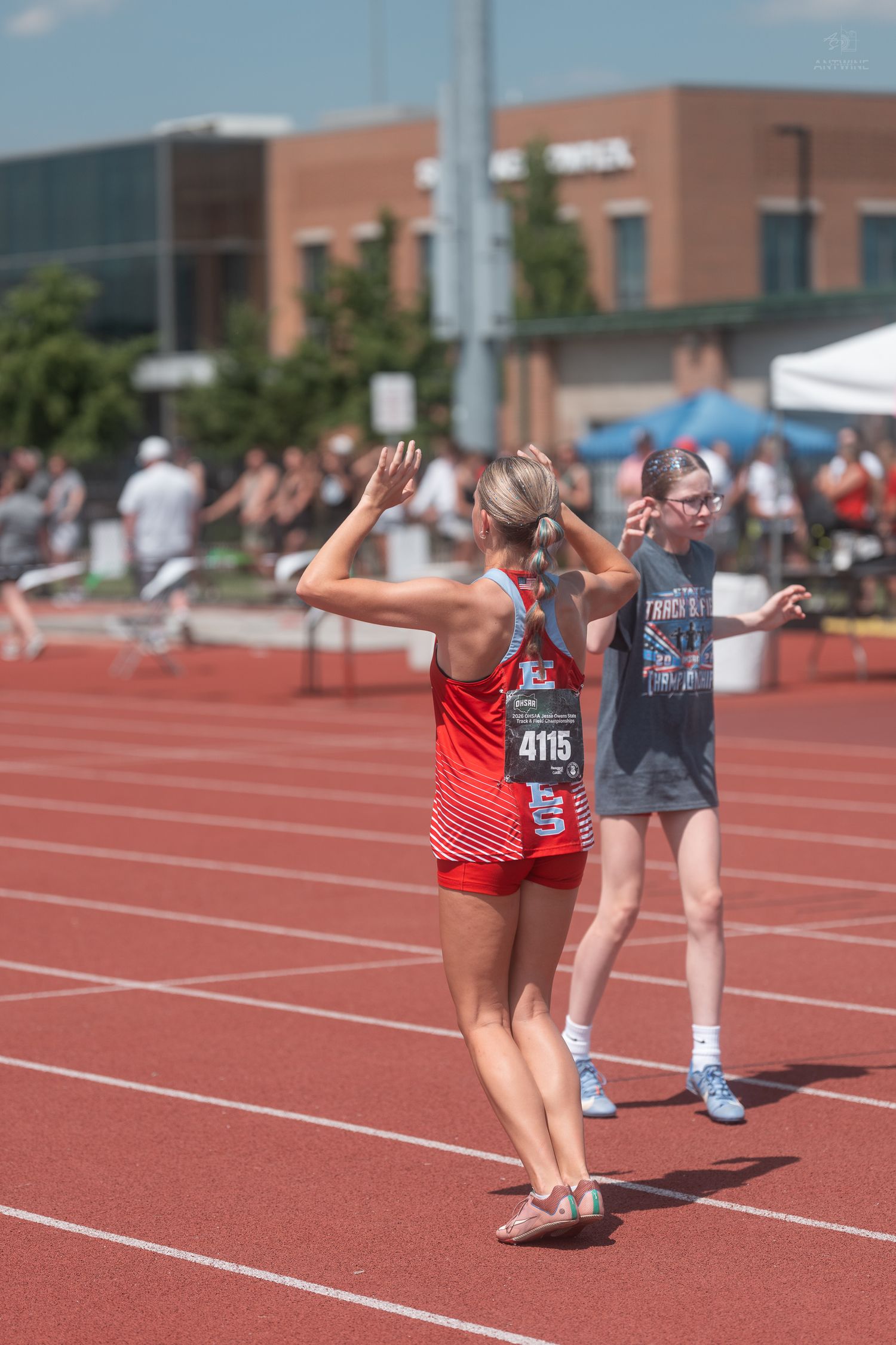 Female track athlete number 4115 in red Eastwind uniform raises hands on red track at outdoor competition.