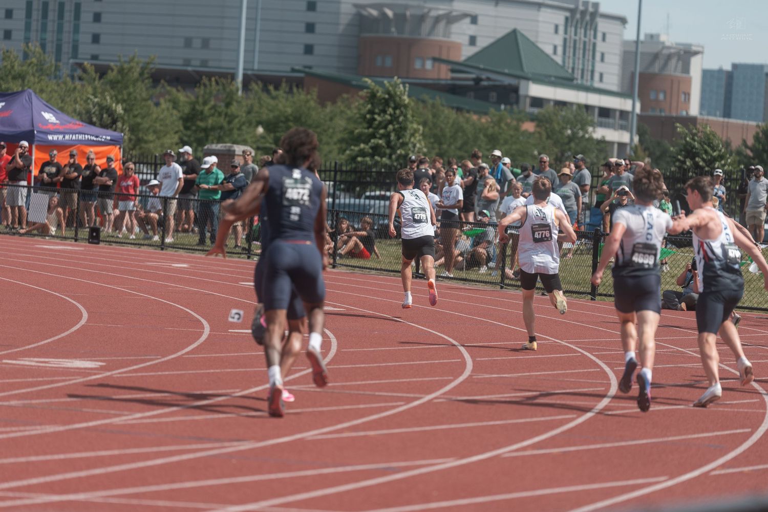 Male track athletes sprint toward finish line during competitive outdoor relay race event.
