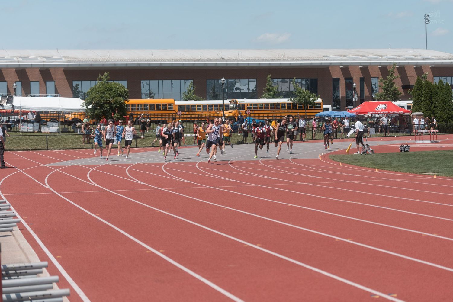 Athletes compete in a running event on a red outdoor track with school buses and a stadium in the background.