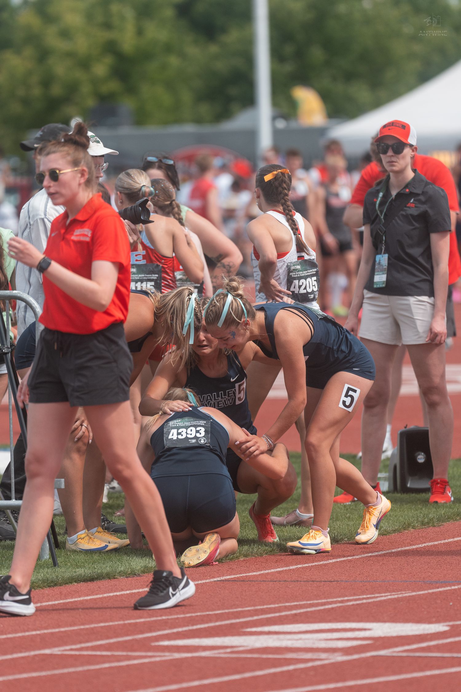 Female athlete wearing bib 4393 kneels on the track while teammates comfort her after a relay race.