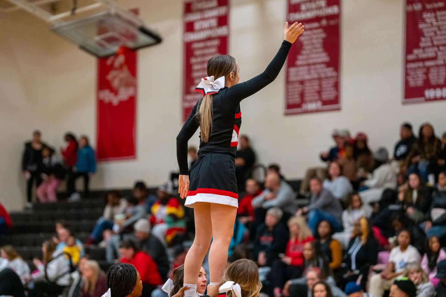 High school cheerleader stands elevated in a stunt formation waving to the crowd during a basketball game.