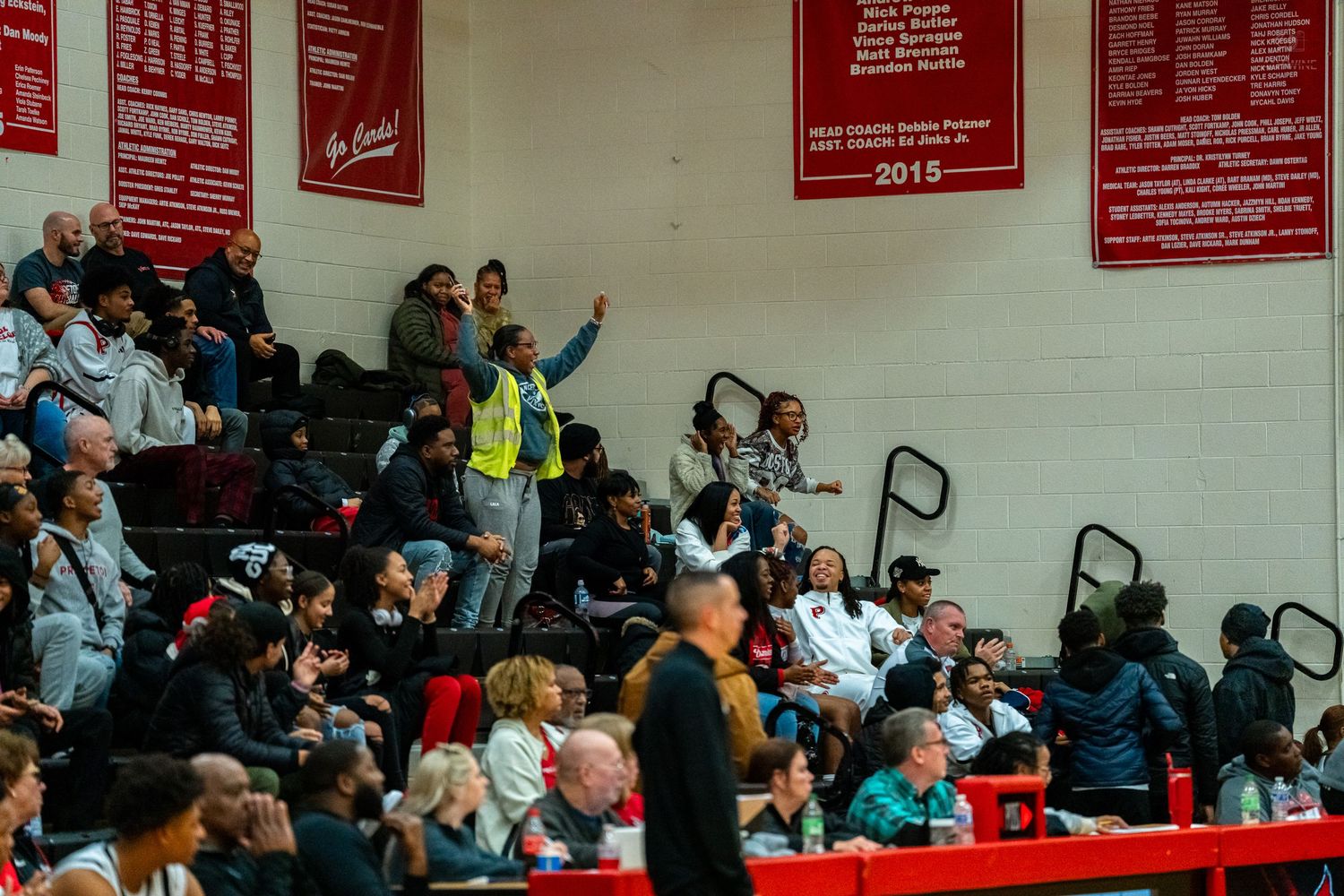 Crowded high school basketball game bleachers with fans cheering, red championship banners visible on gymnasium wall.