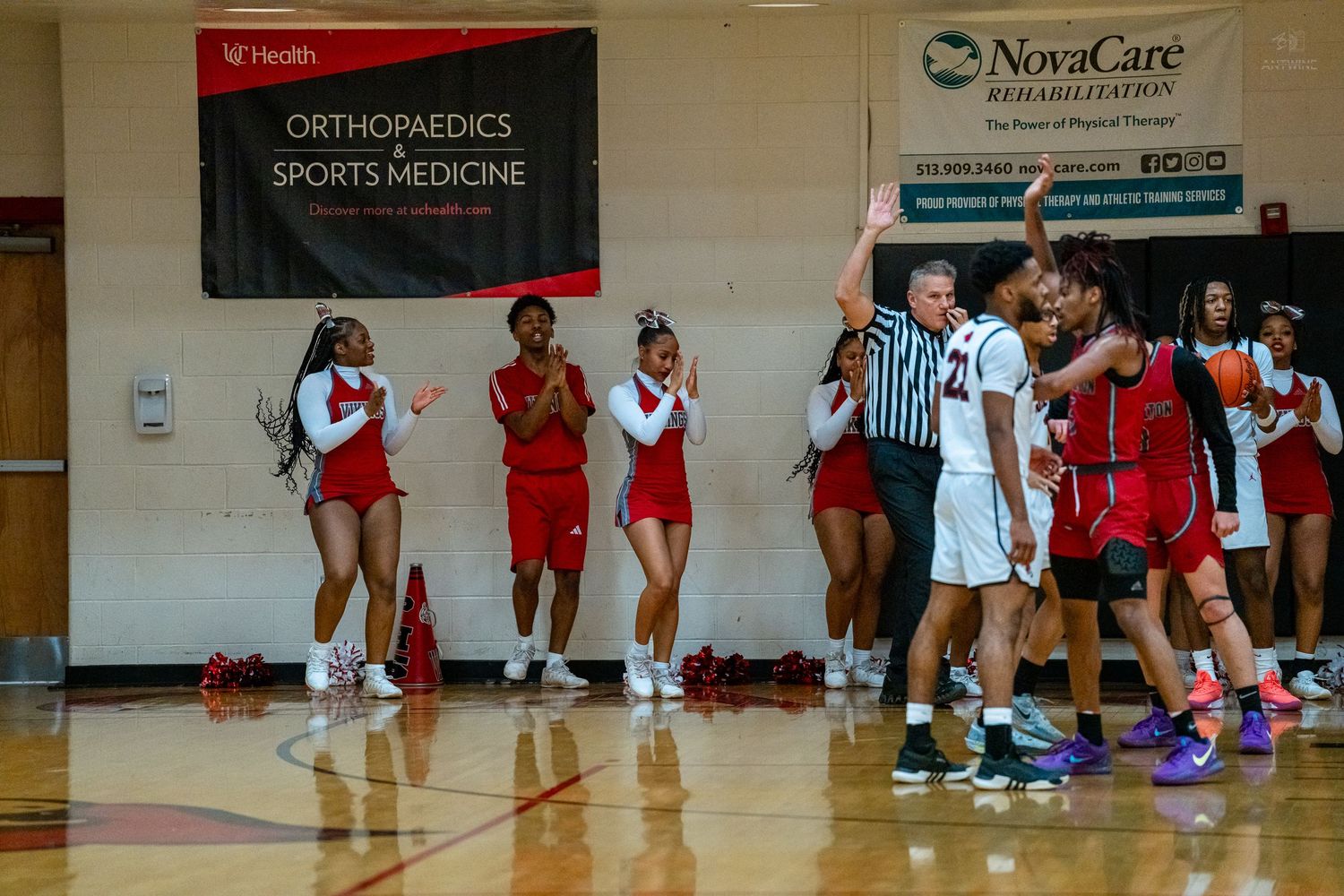 Cheerleaders in red and white uniforms cheer on the sideline during a high school basketball game in a gymnasium.