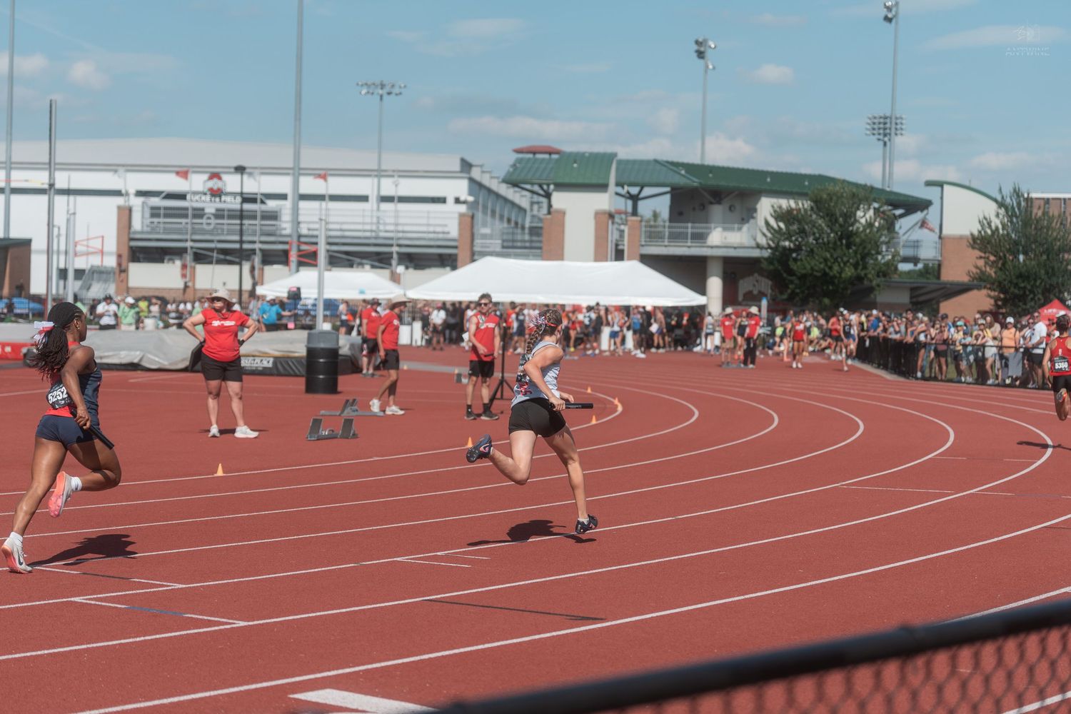 Female relay runners exchange a baton on a red track at an outdoor track and field competition.