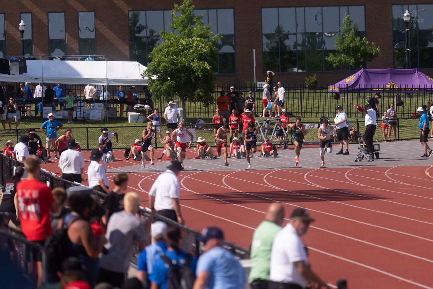 Runners and spectators fill a red track during an outdoor high school track and field meet on a bright sunny day.
