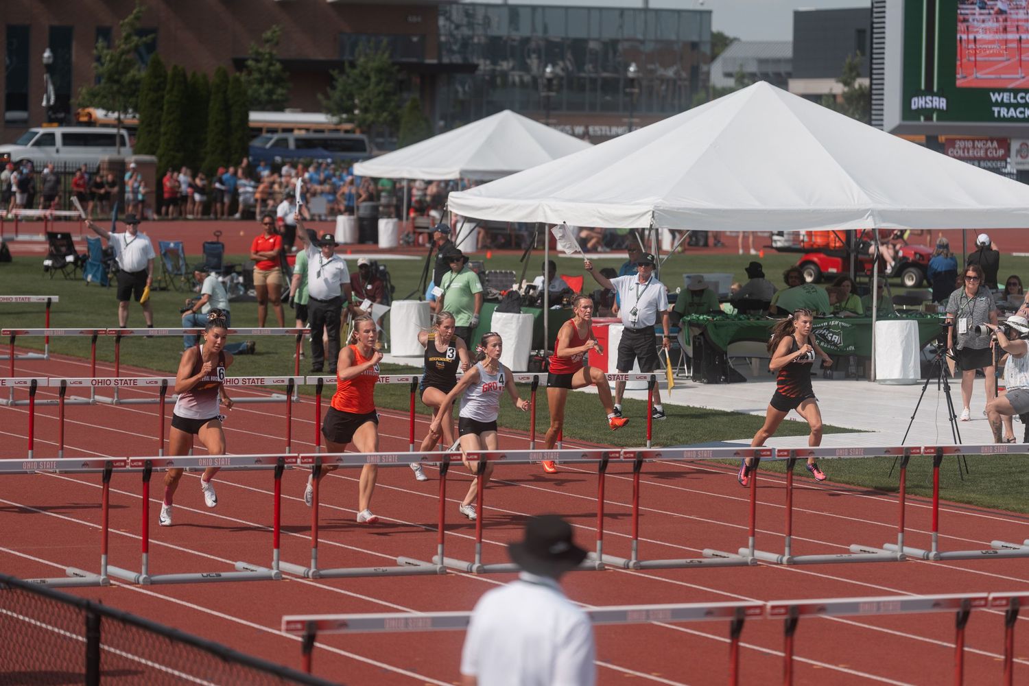 Female athletes compete in hurdles event at outdoor track meet with white tents and crowd visible.