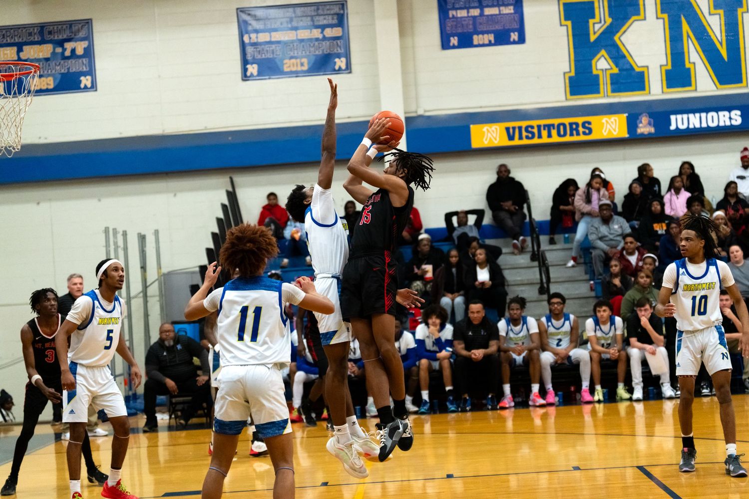 Basketball player in red uniform leaps for a shot over defenders in a packed high school gymnasium.