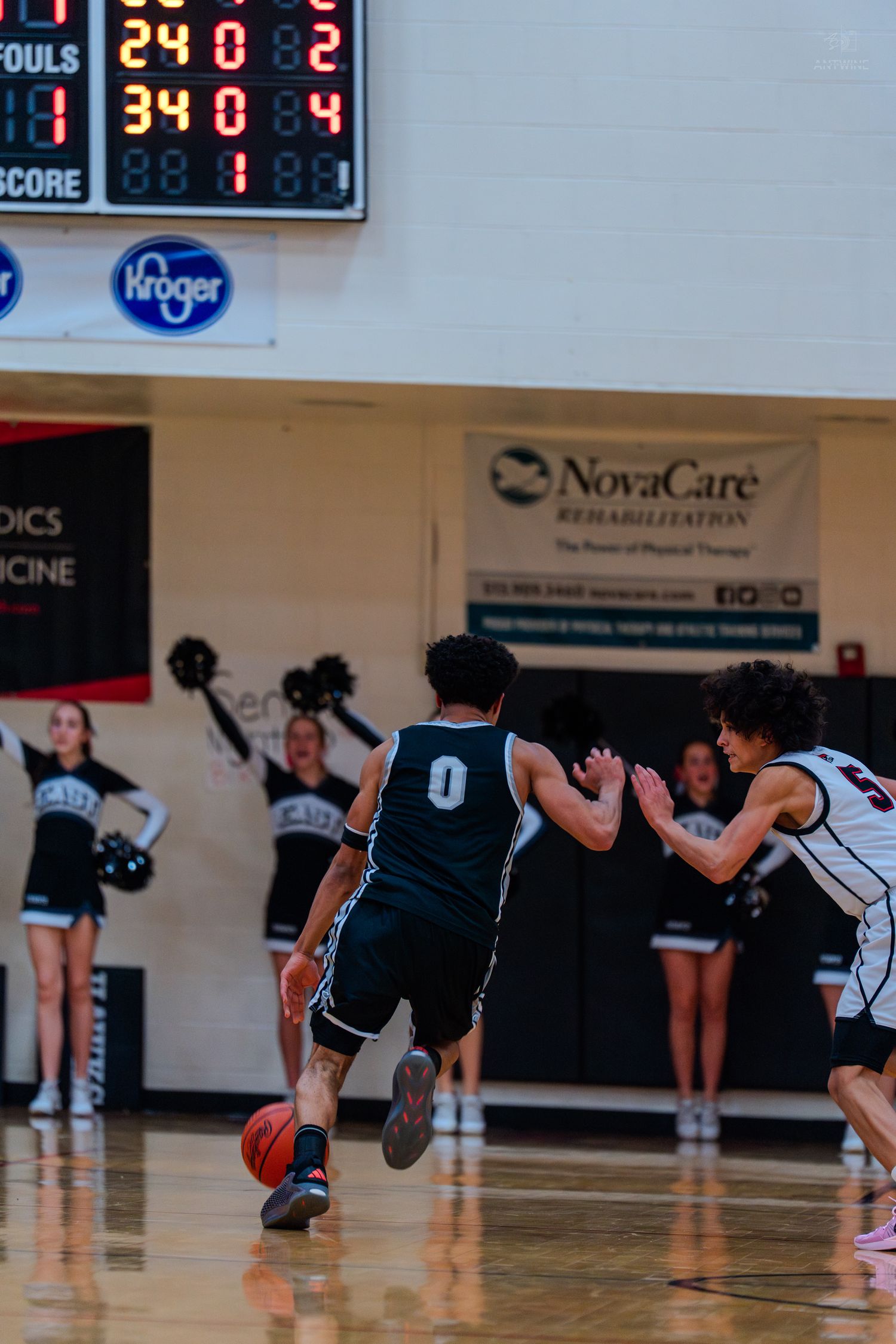 Basketball player #0 drives past defender during a high school game with cheerleaders in background.