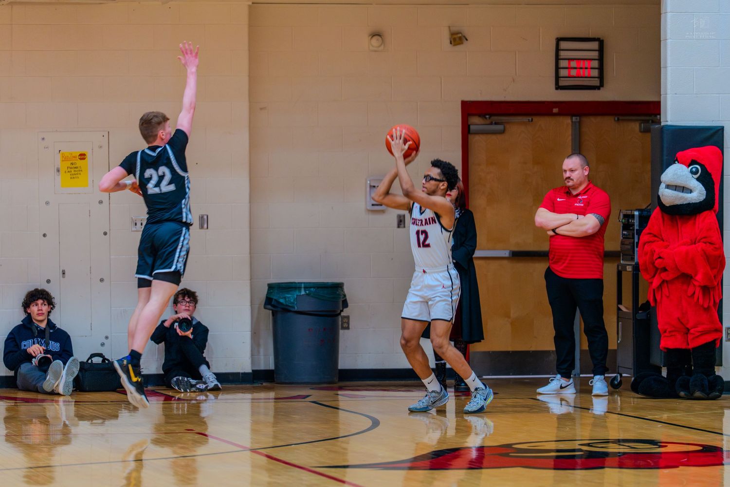 Player #12 shoots over defender #22 during a high school basketball game with a mascot watching courtside.