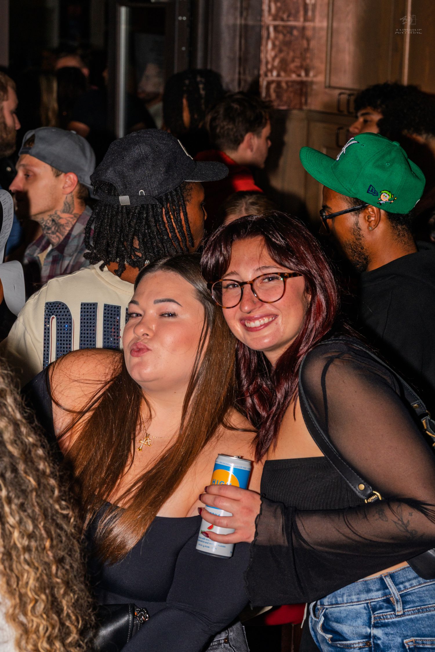 Two women posing together at a lively nightclub, one smiling and holding a canned beverage.