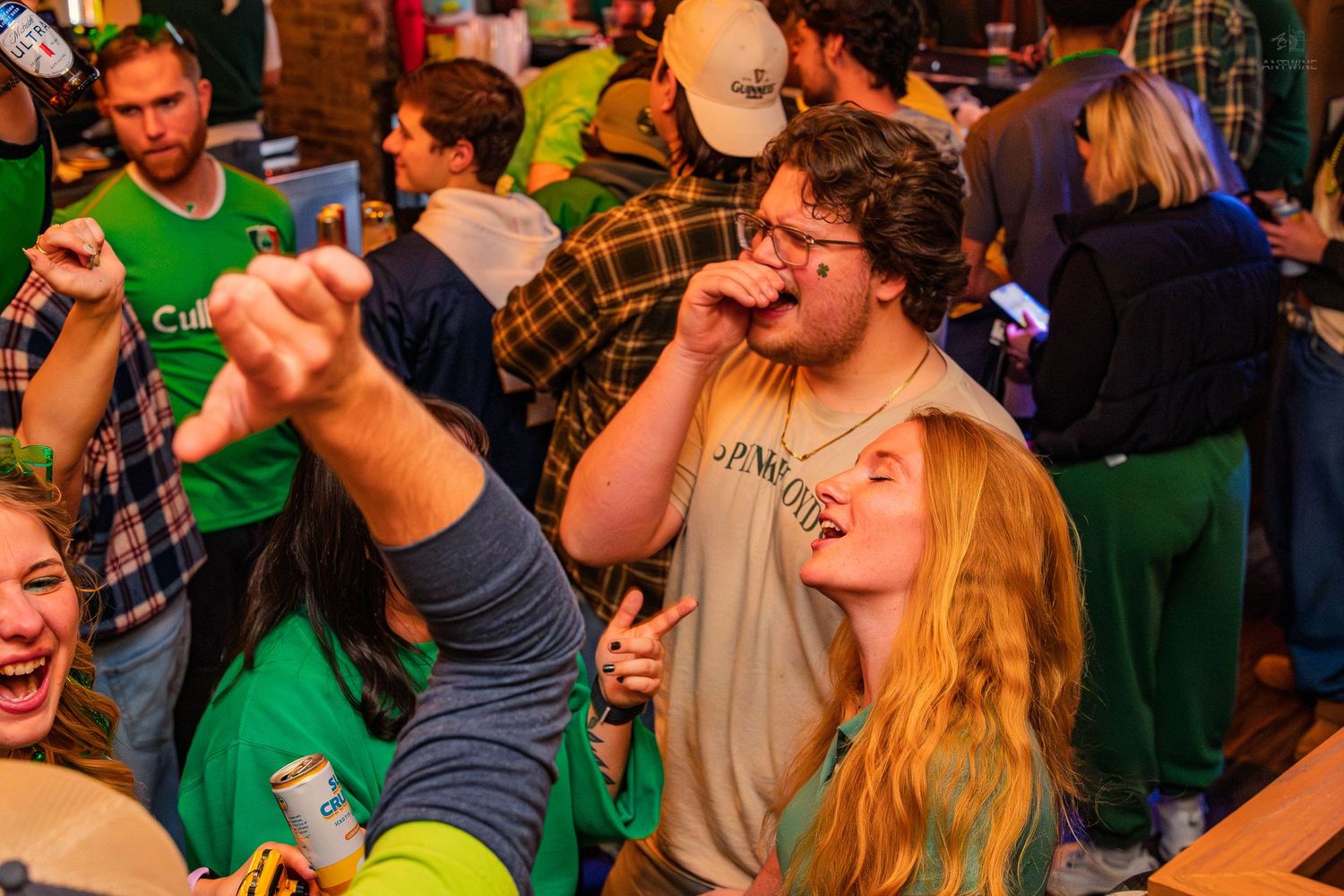 Young couple taking a selfie at a lively St. Patrick's Day bar party with drinks in hand.