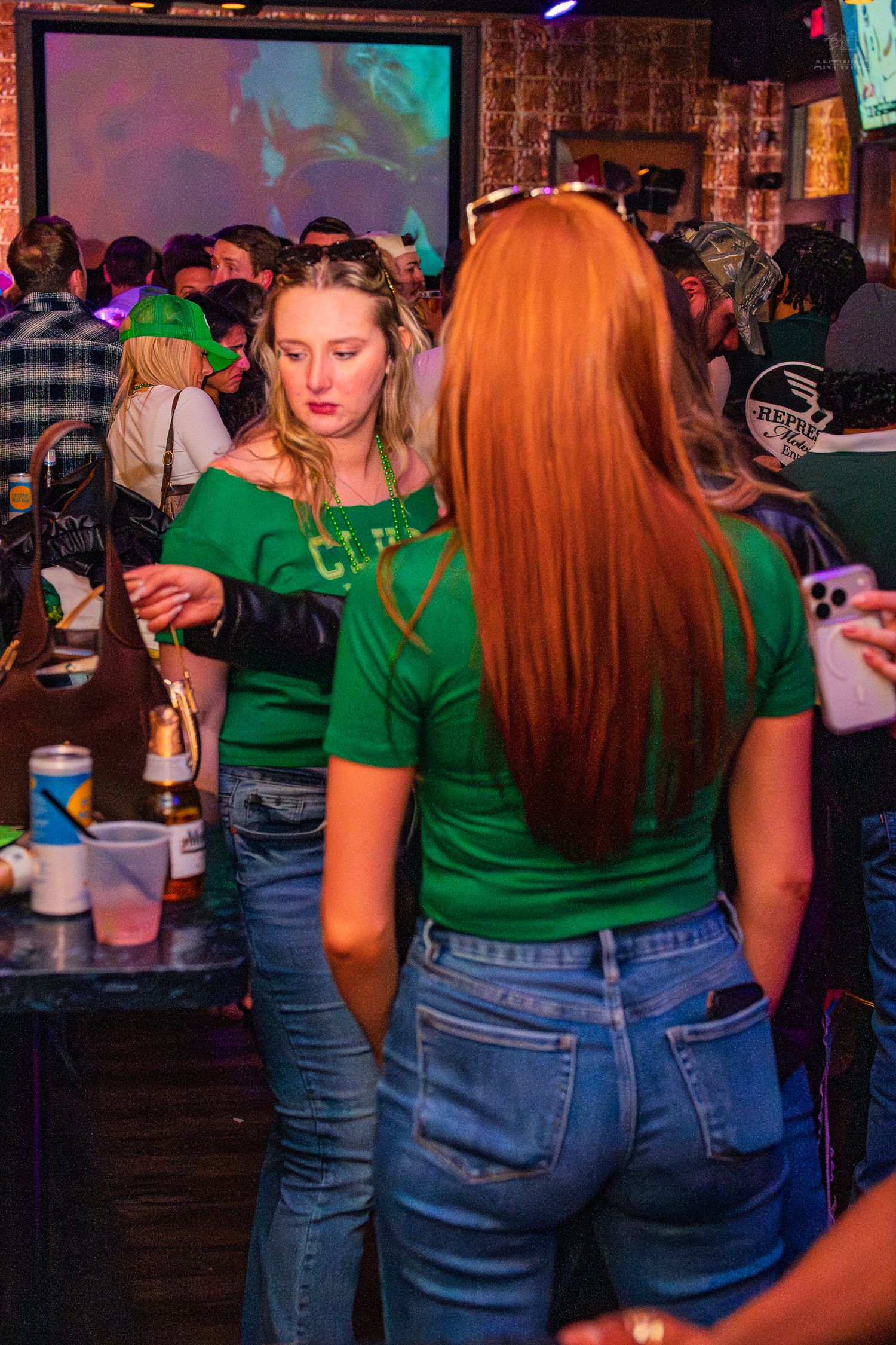 Two women in green shirts chat at a lively St. Patrick's Day bar celebration.