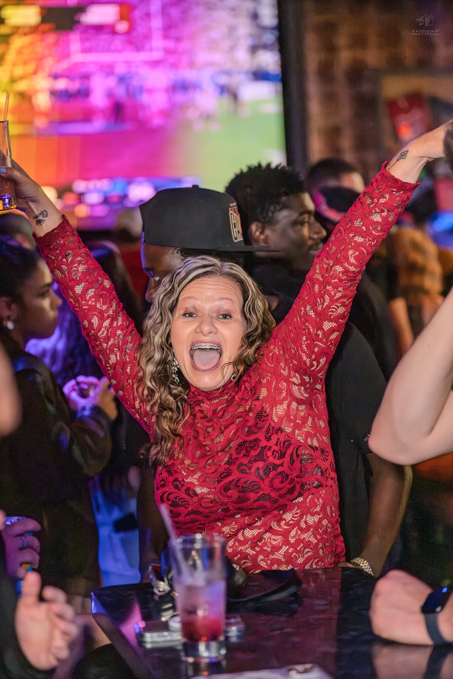 Enthusiastic woman in red lace dress raises arms with tongue out at a crowded nightclub party.
