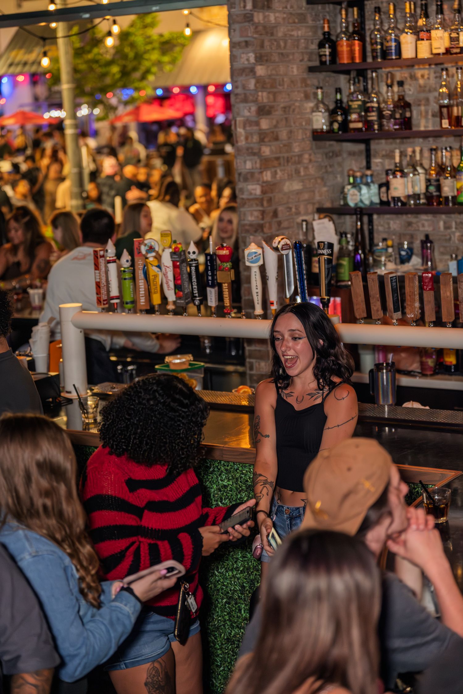 Bartender in black top and green skirt serving customers at a busy bar with draft beer taps.