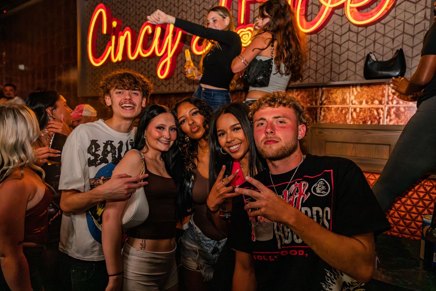 Group of young friends posing together at a fun nightclub with neon Cincinnati sign.