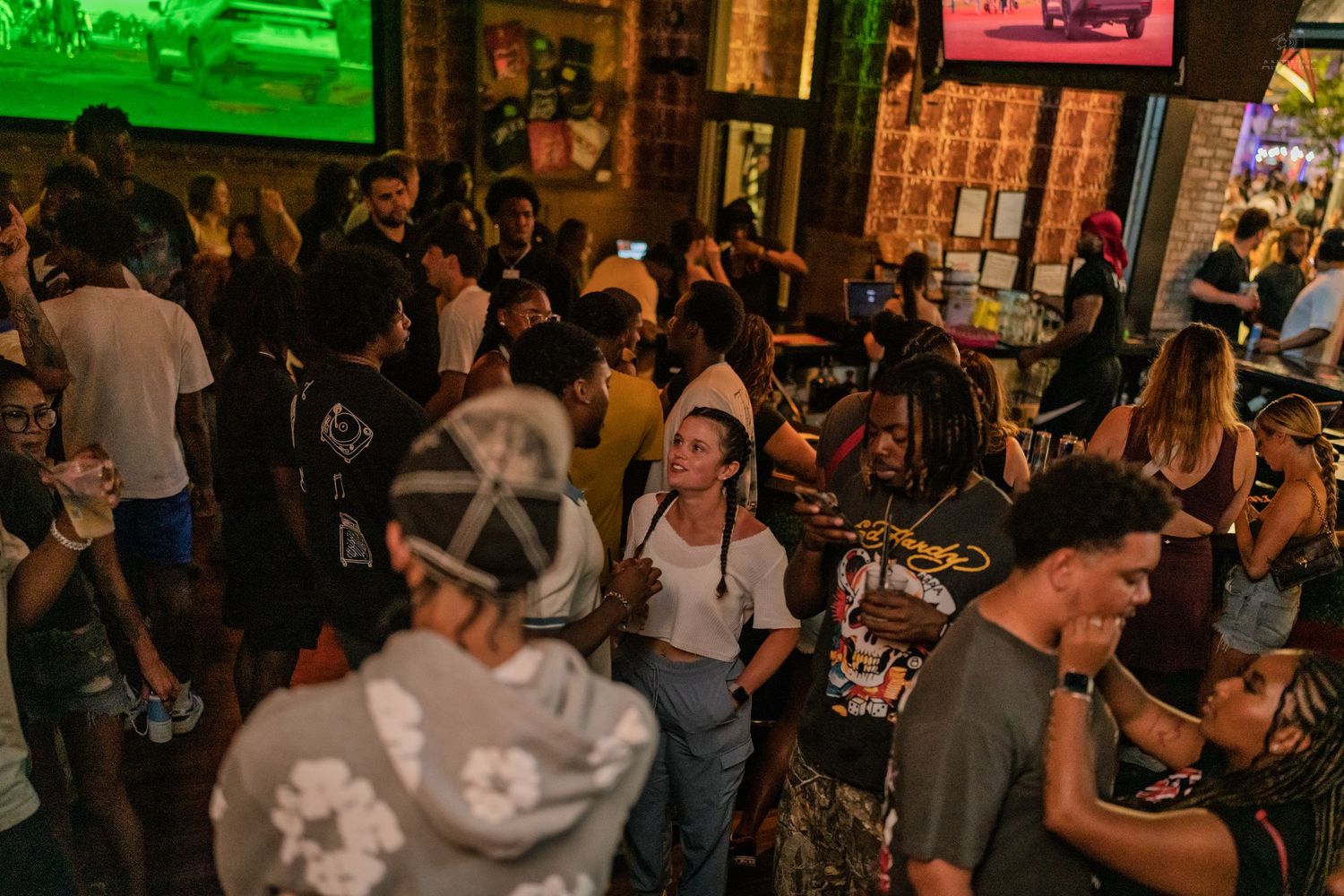Diverse crowd of people socializing at a lively bar with green neon lights and TVs.