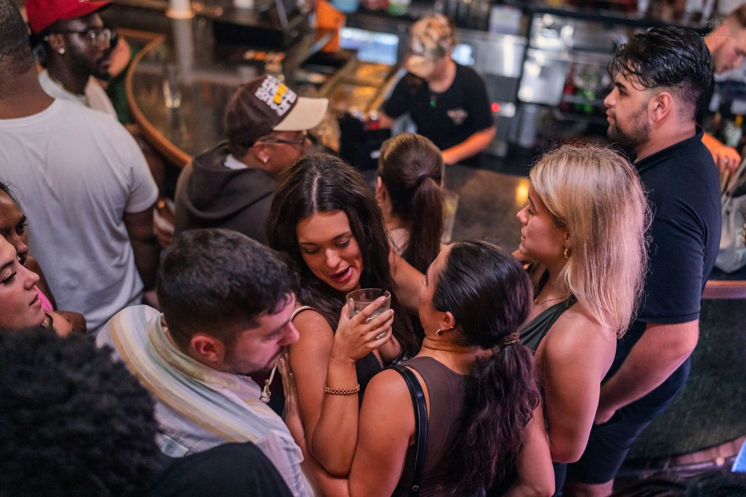 Group of young women laughing and hugging together near a bar in a busy nightclub.