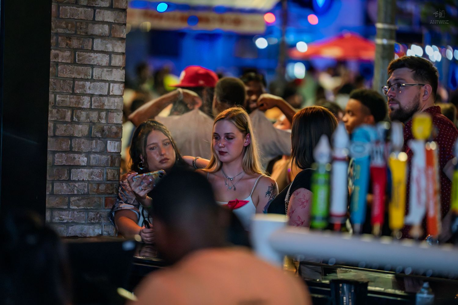 Two women leaning against a brick pillar near a bar with colorful tap handles at night.