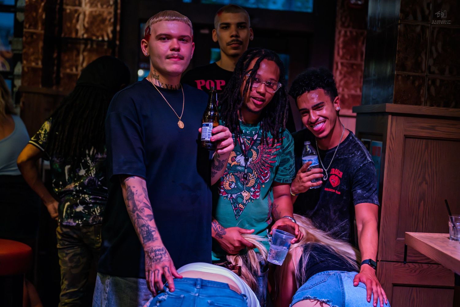 Three young men posing together at a bar, holding drinks in a casual nightlife setting.