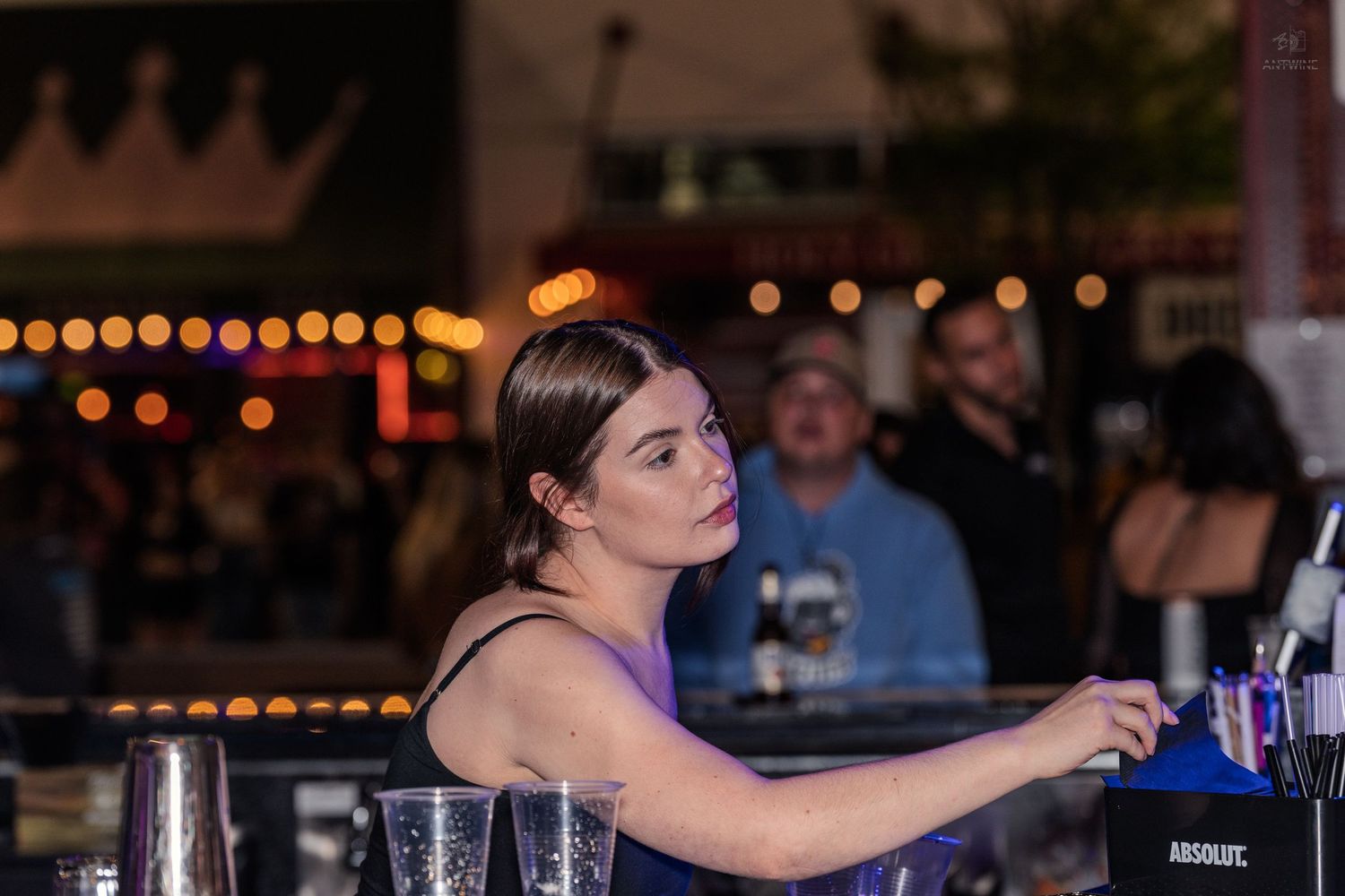 Female bartender working behind a bar with Absolut vodka bottles at an outdoor night event.