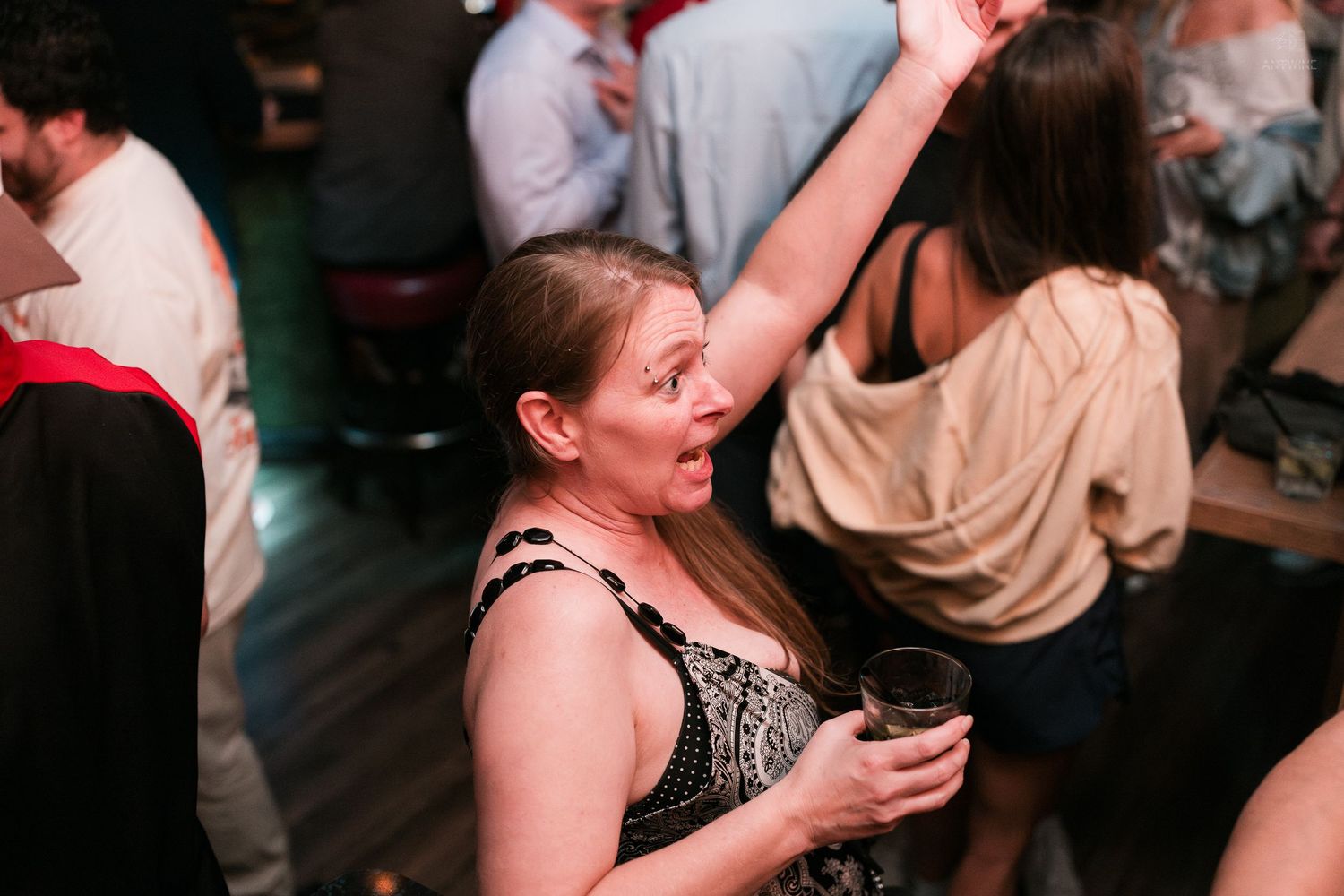 An excited woman raises her arm while holding a drink on a crowded nightclub dance floor.
