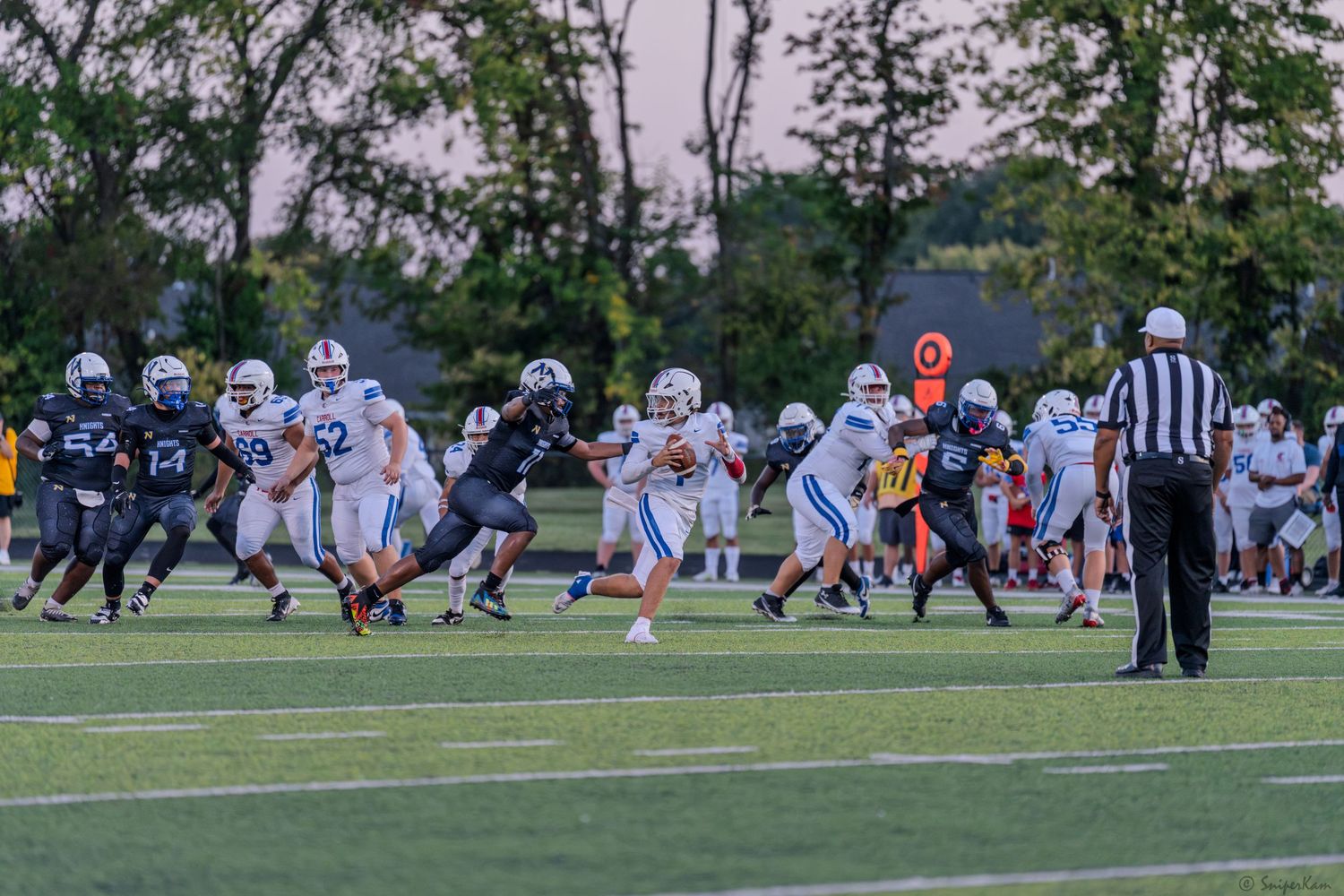 High school football players in blue and black uniforms clash during an evening game with a referee nearby.