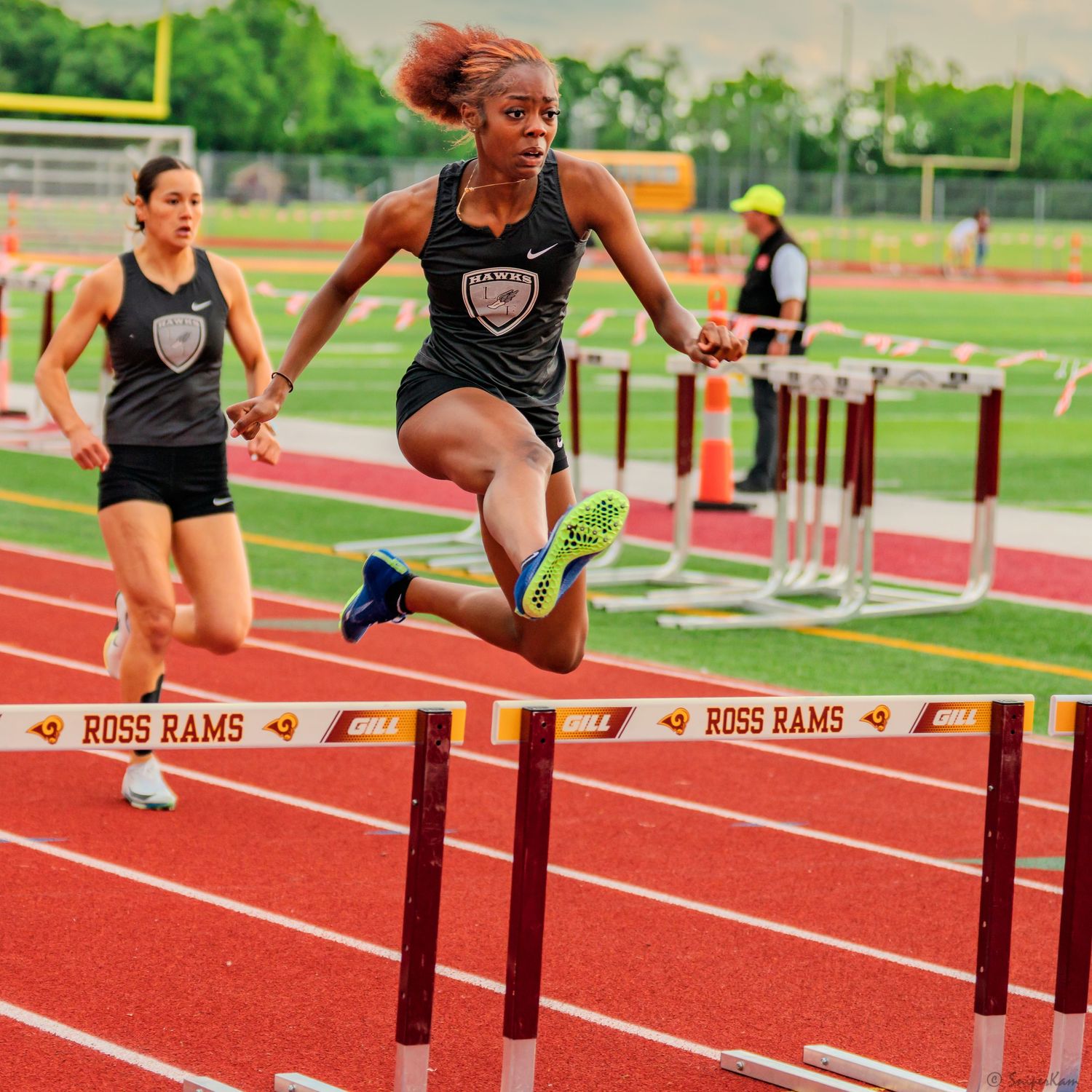 Female athlete powerfully clears a hurdle on a Ross Rams track during a competitive meet.