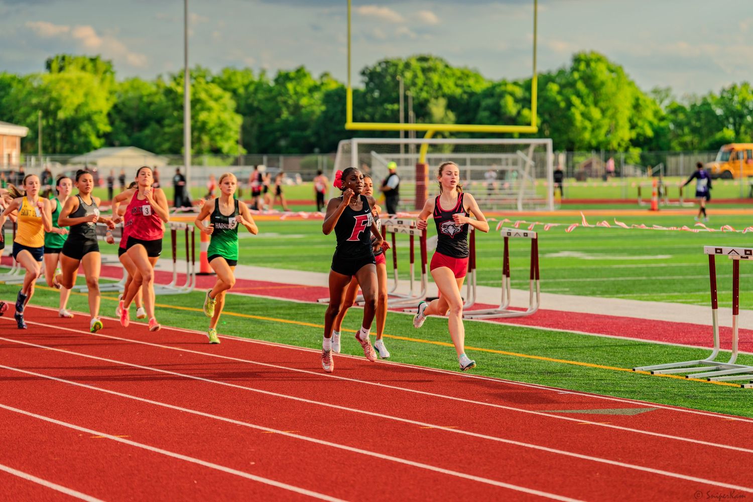 Female runners compete in a track race with hurdles at outdoor stadium.