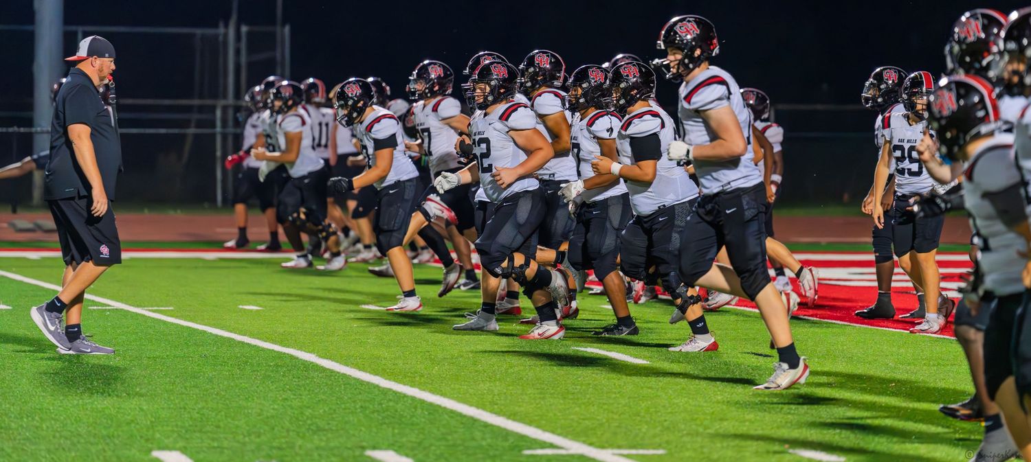 High school football team in dark uniforms jogs onto the field during a night game warm-up.