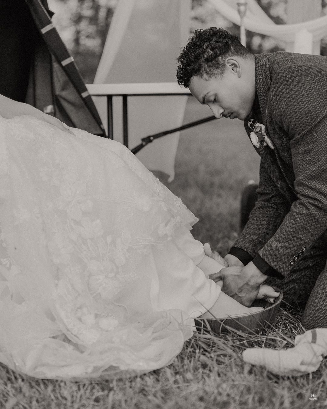 Black and white photo of a groom kneeling to adjust the bride's wedding dress hem on grass.