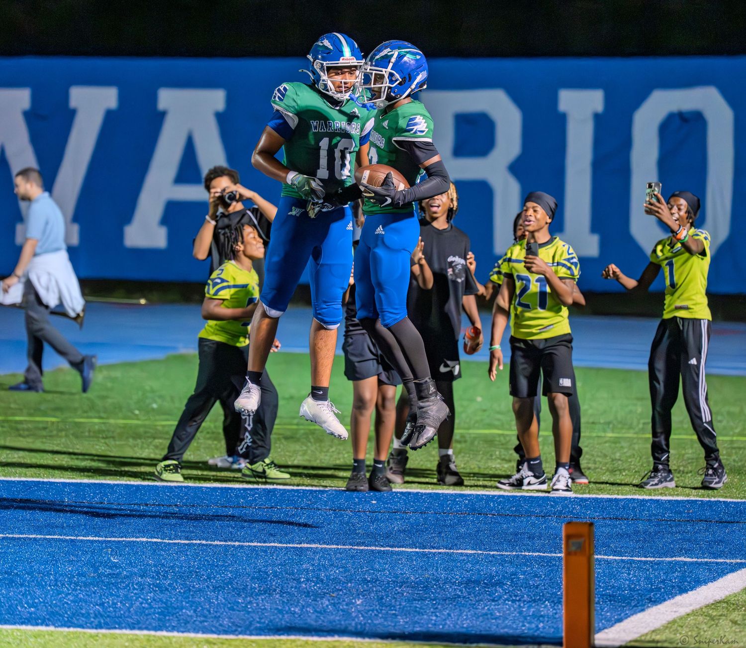 Warriors football players in green and blue uniforms celebrate a touchdown in the end zone at night.