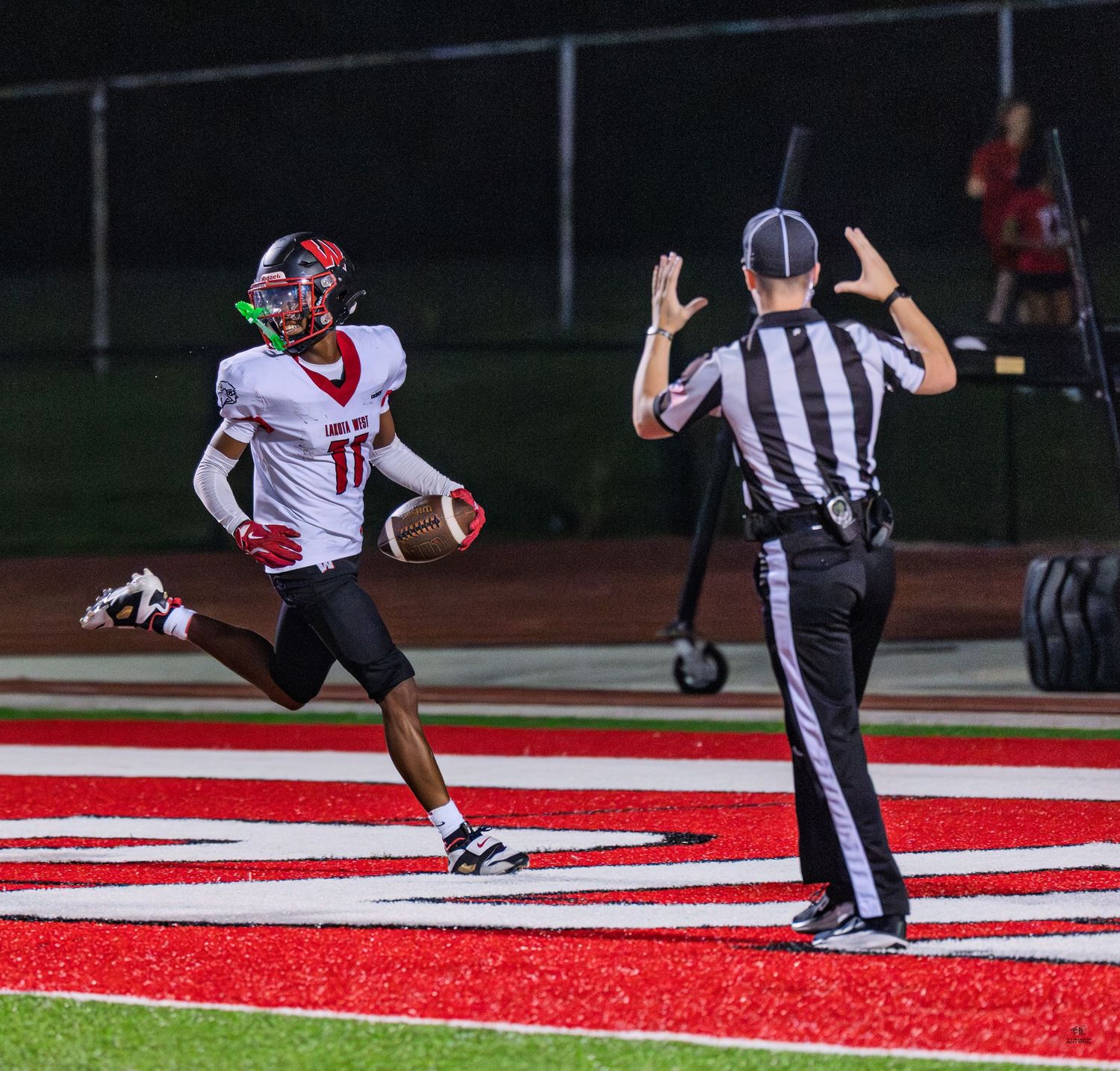Player number 11 celebrates in red end zone as referee signals touchdown during high school football game.