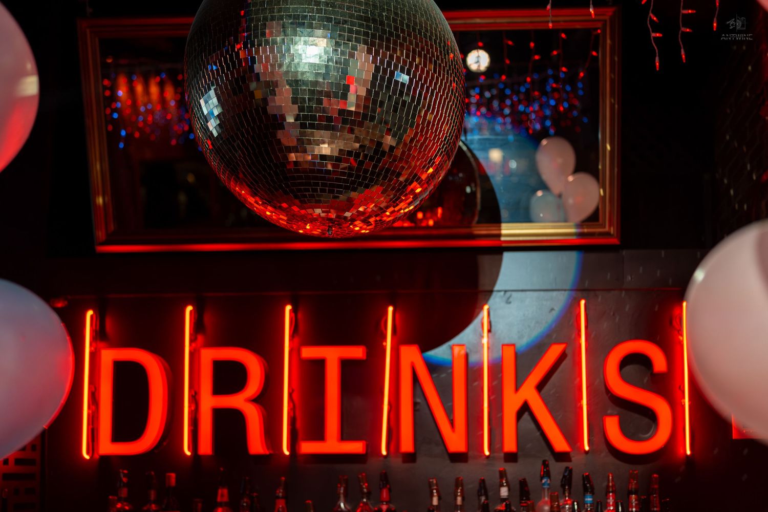 Neon red DRINKS sign glowing below a disco ball in a vibrant nightclub bar setting with balloons.