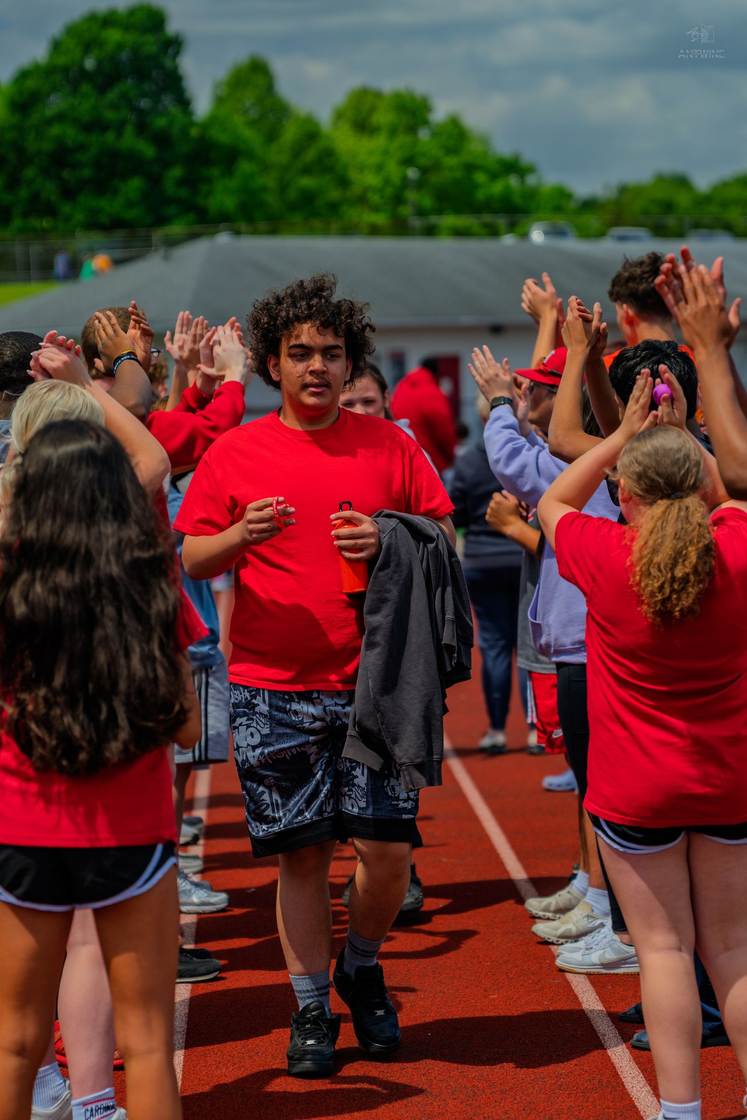 Students in red shirts form a celebratory tunnel on a track, cheering for a curly-haired teen walking through.
