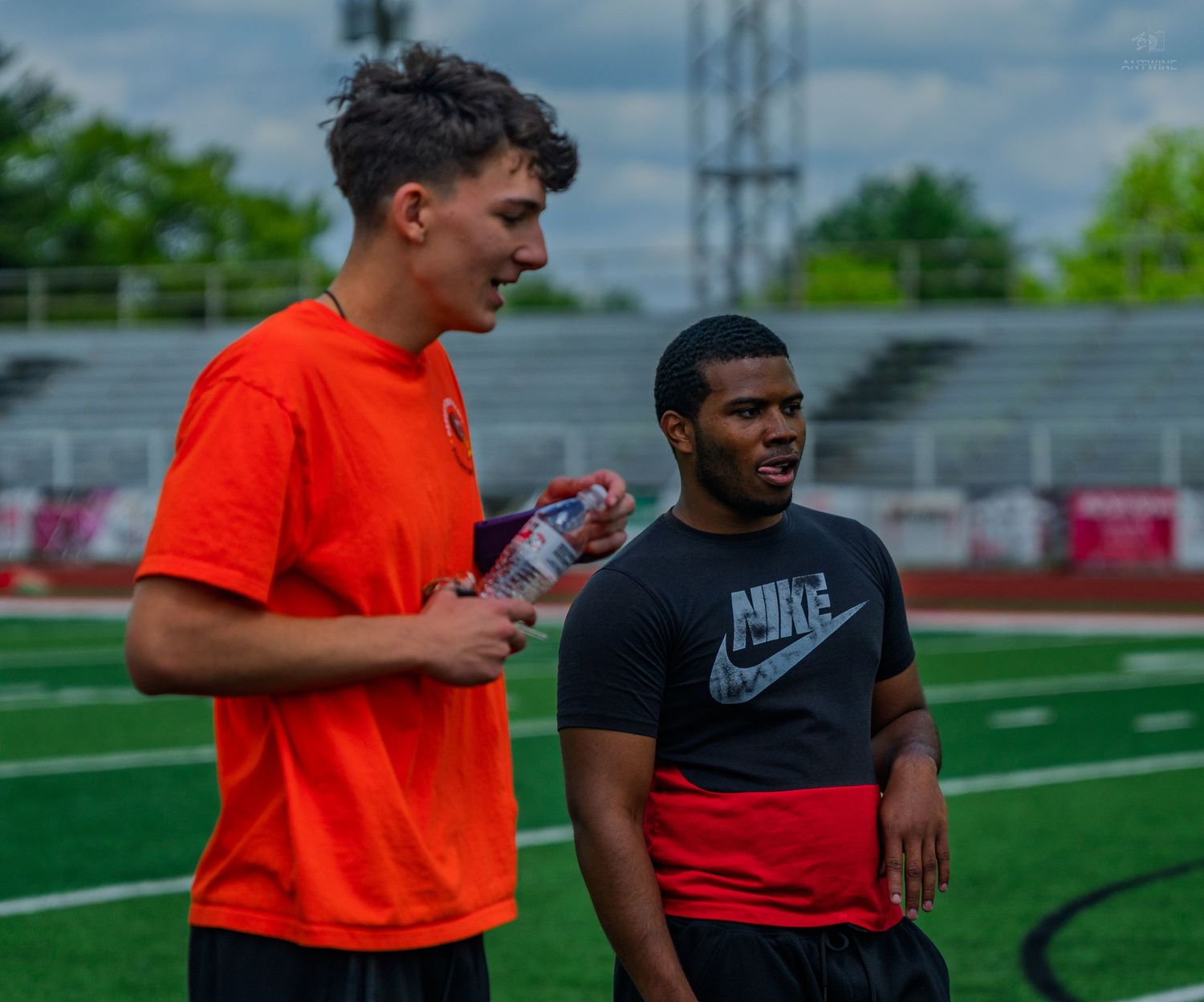 Two young athletes in Nike apparel chat on a football field, one holding a water bottle.