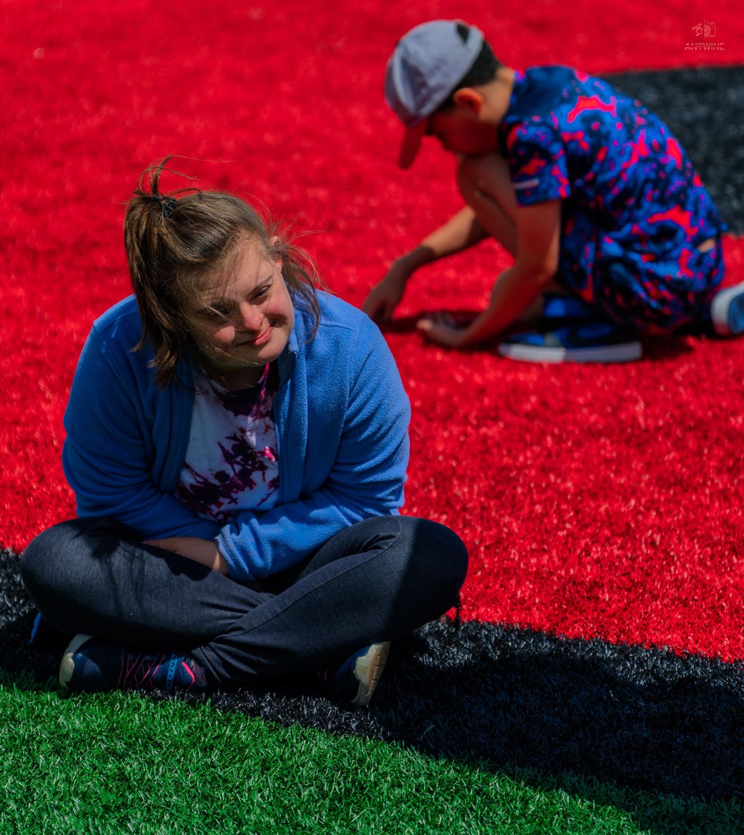 Two people sitting on vibrant red and green turf outdoors, woman in blue hoodie cross-legged in foreground.