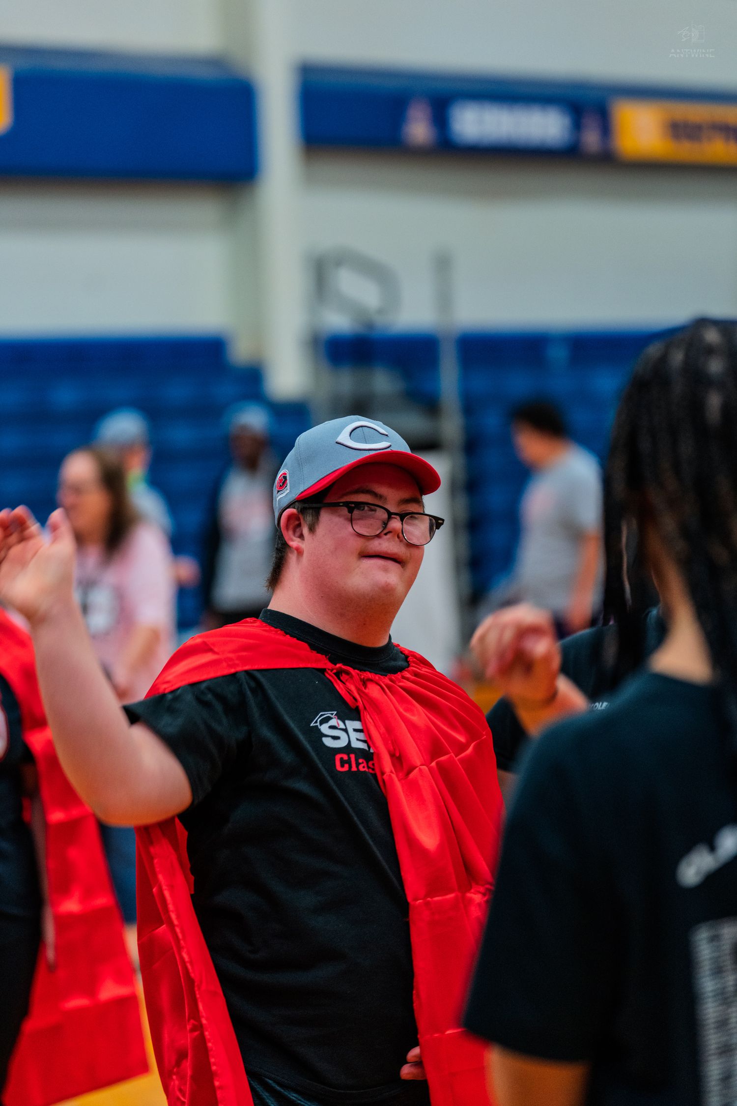 Young man with Down syndrome smiling in red cape and cap at a Special Olympics basketball event.