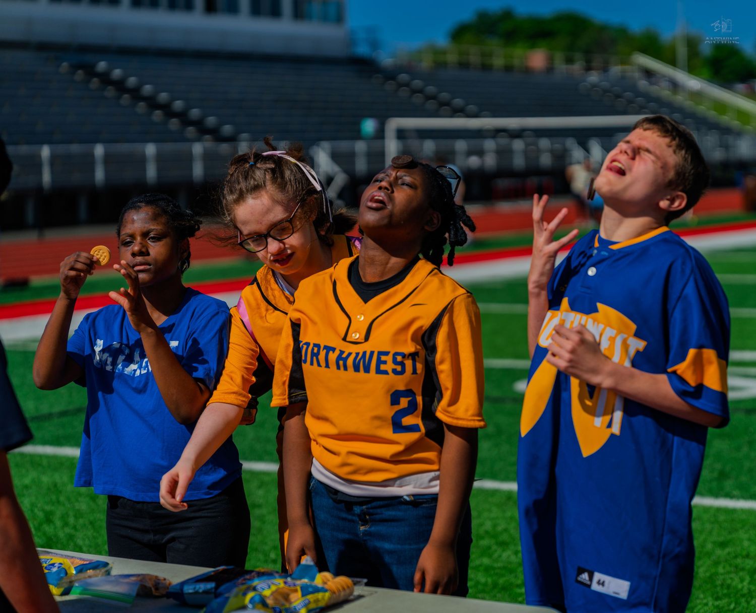 Kids in sports jerseys laugh and play outdoors near a stadium field on a sunny day.