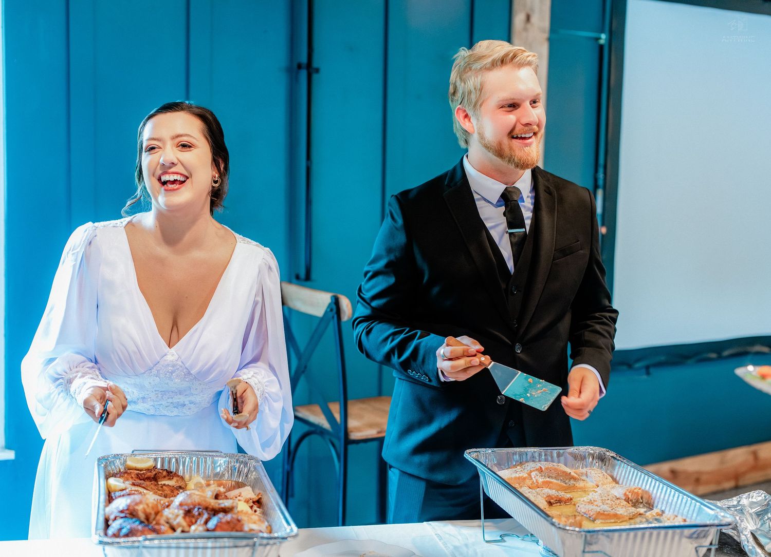 Bride and groom laughing together at wedding reception buffet table with food trays, bride in white gown, groom in black suit.