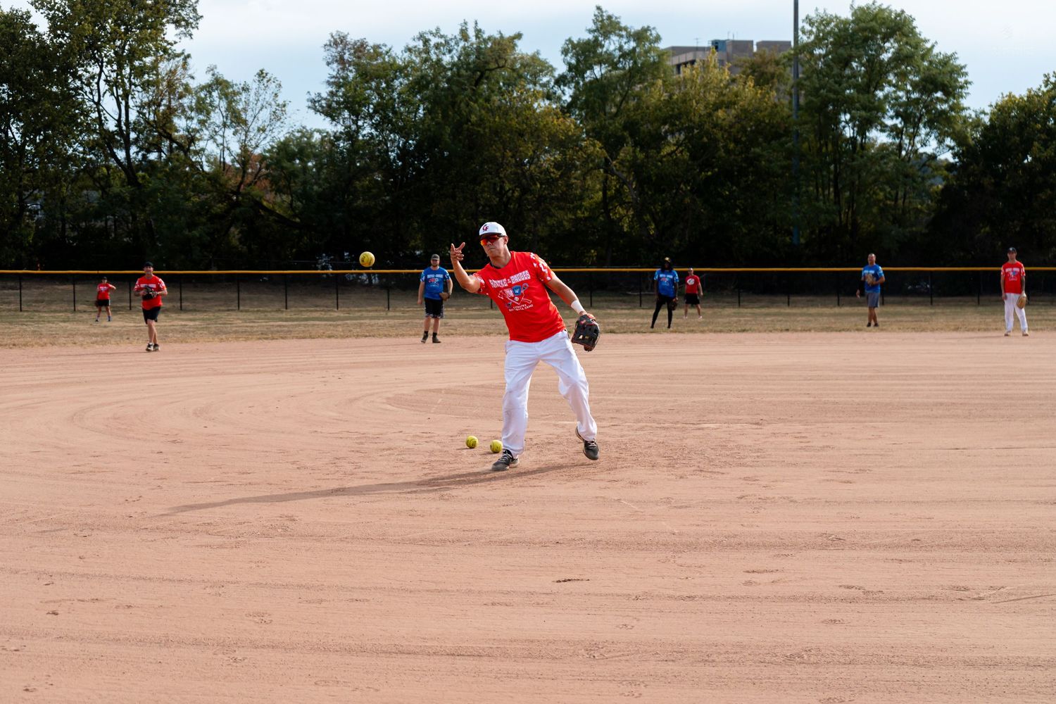 Player in red jersey throws ball on dirt softball field during outdoor recreational game with teammates in background.