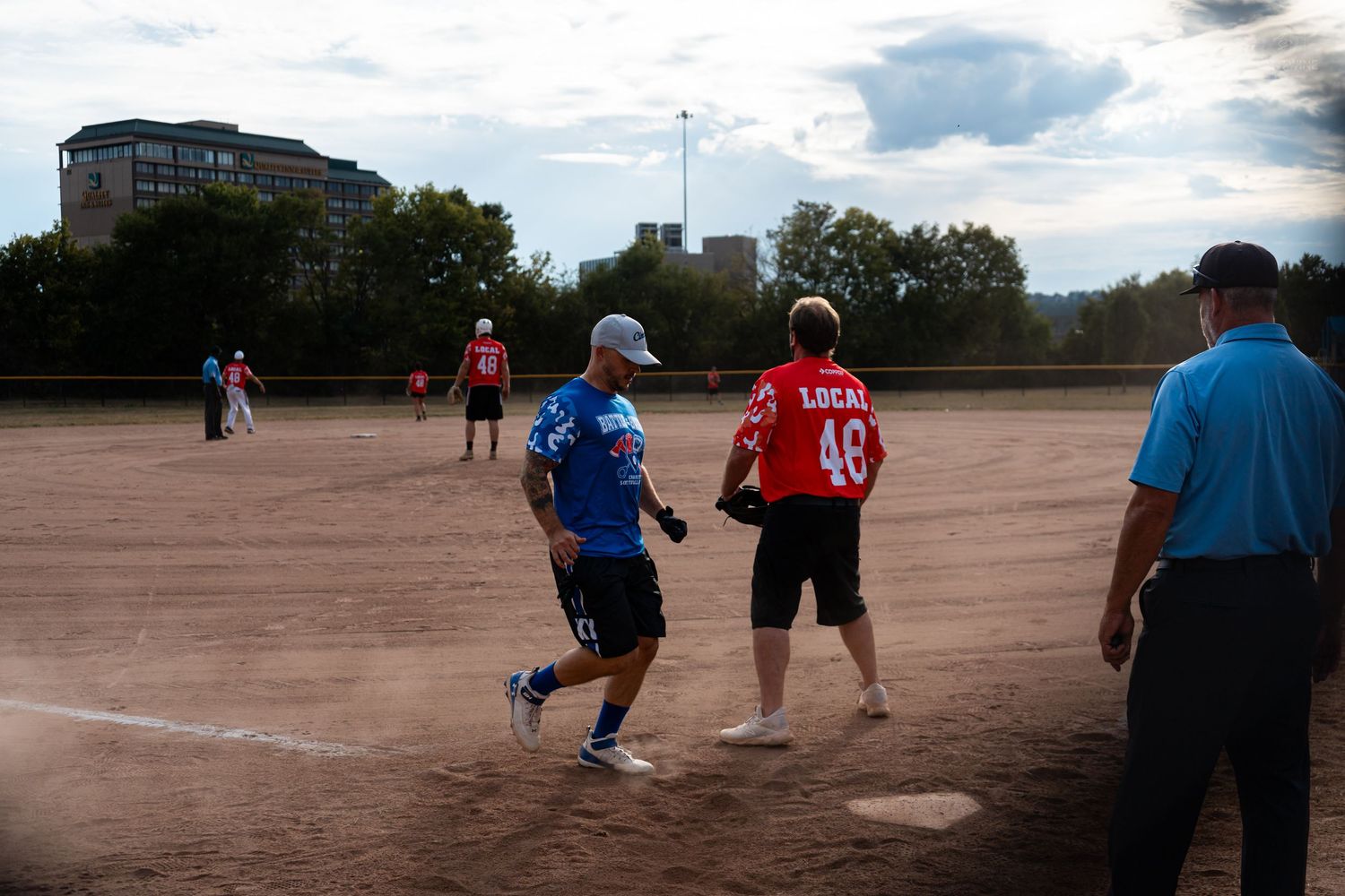 Softball players in blue and red Local 48 jerseys round the bases during an evening recreational league game.