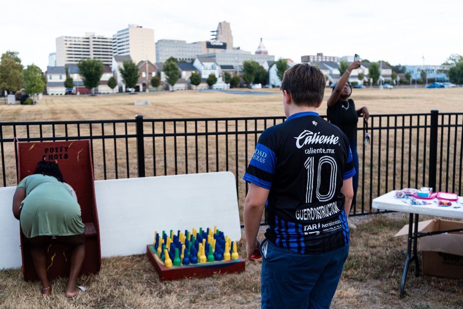 A person in a Chivas jersey watches lawn games set up near a softball field with city skyline views.