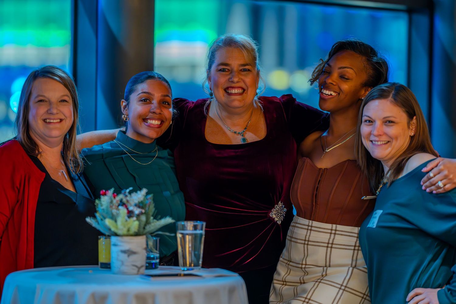Five women smiling and posing together at an indoor event, standing around a small table with a centerpiece.