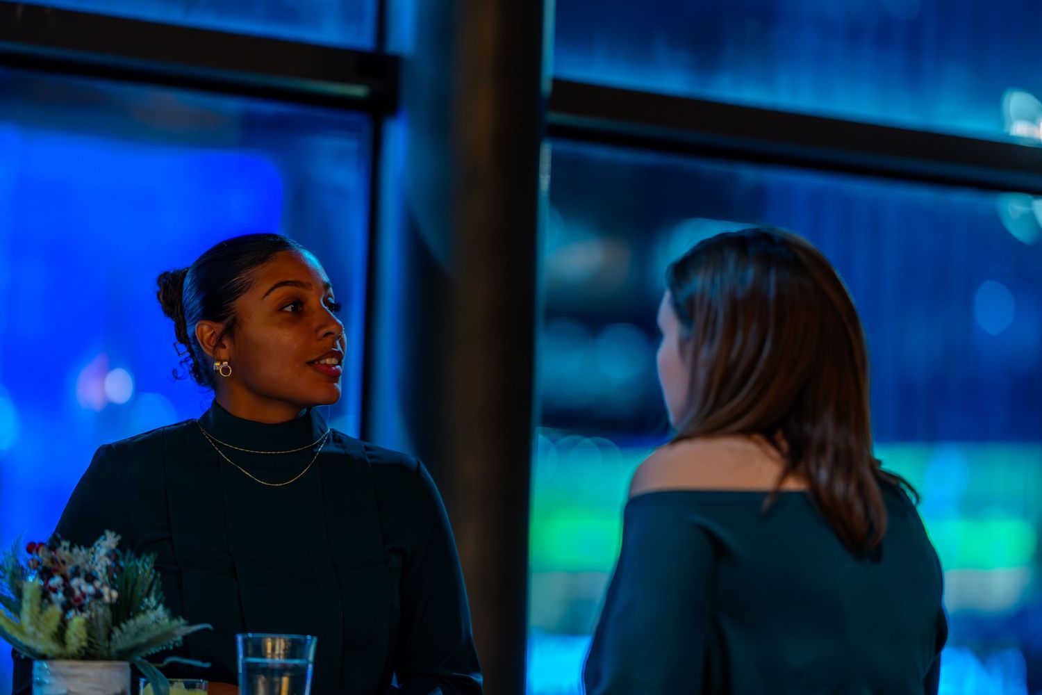 Two women in teal tops engage in conversation near blue-lit windows at an evening social event.