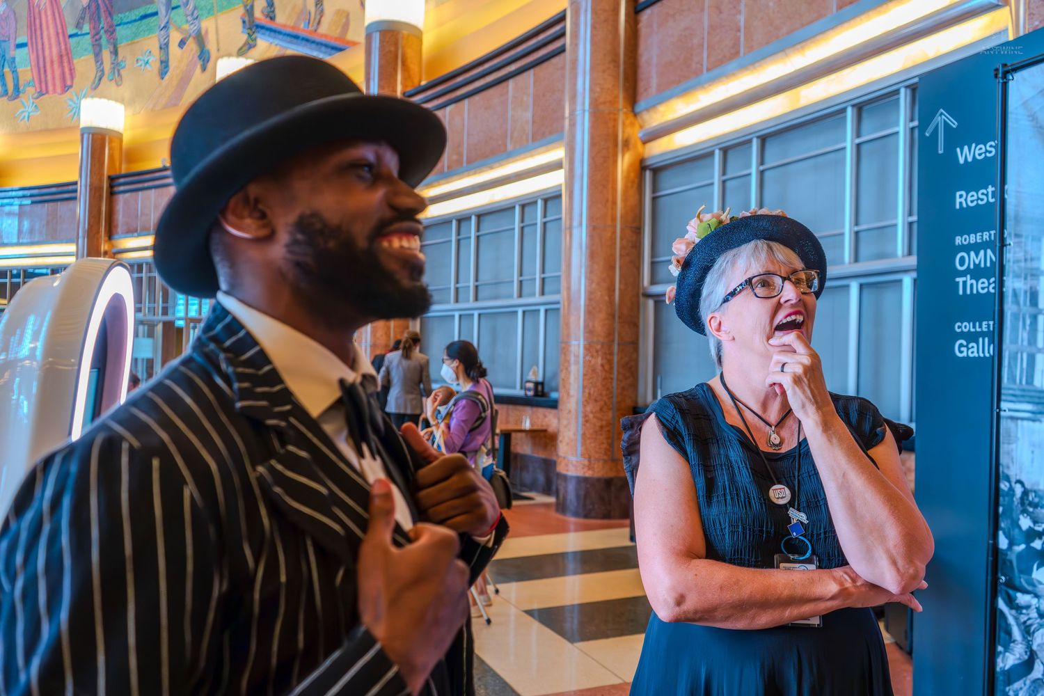 A man in a pinstripe suit and fedora hat chats with a woman in a blue dress at an indoor venue.